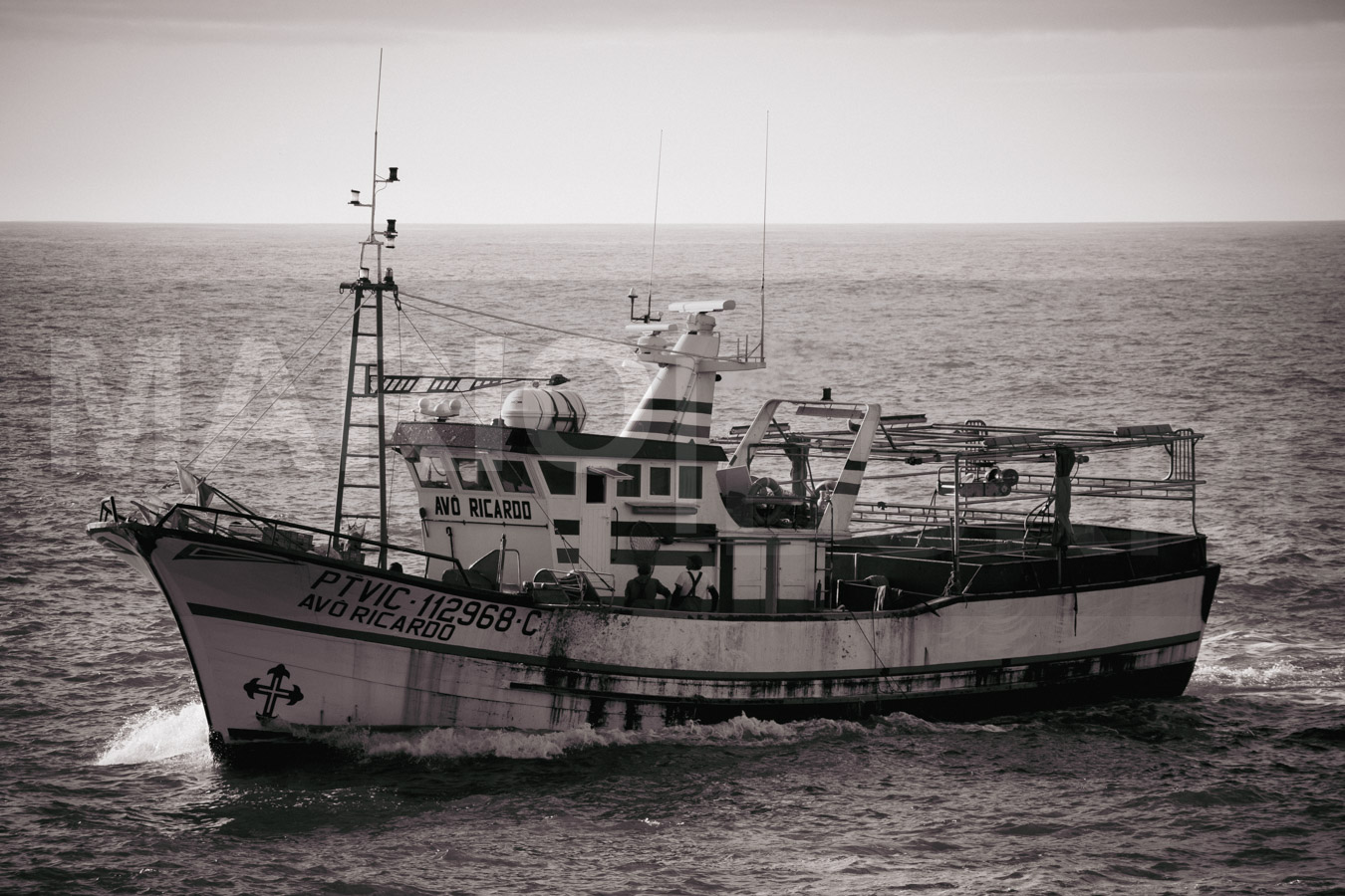 Ein robustes Fischerboot namens "AVÔ RICARDO" gleitet auf einem unruhigen Meer und erzeugt eine Bugwelle unter einem bewölkten Himmel, dargestellt in Schwarz-Weiss. Foto von MARIO|BURRI.