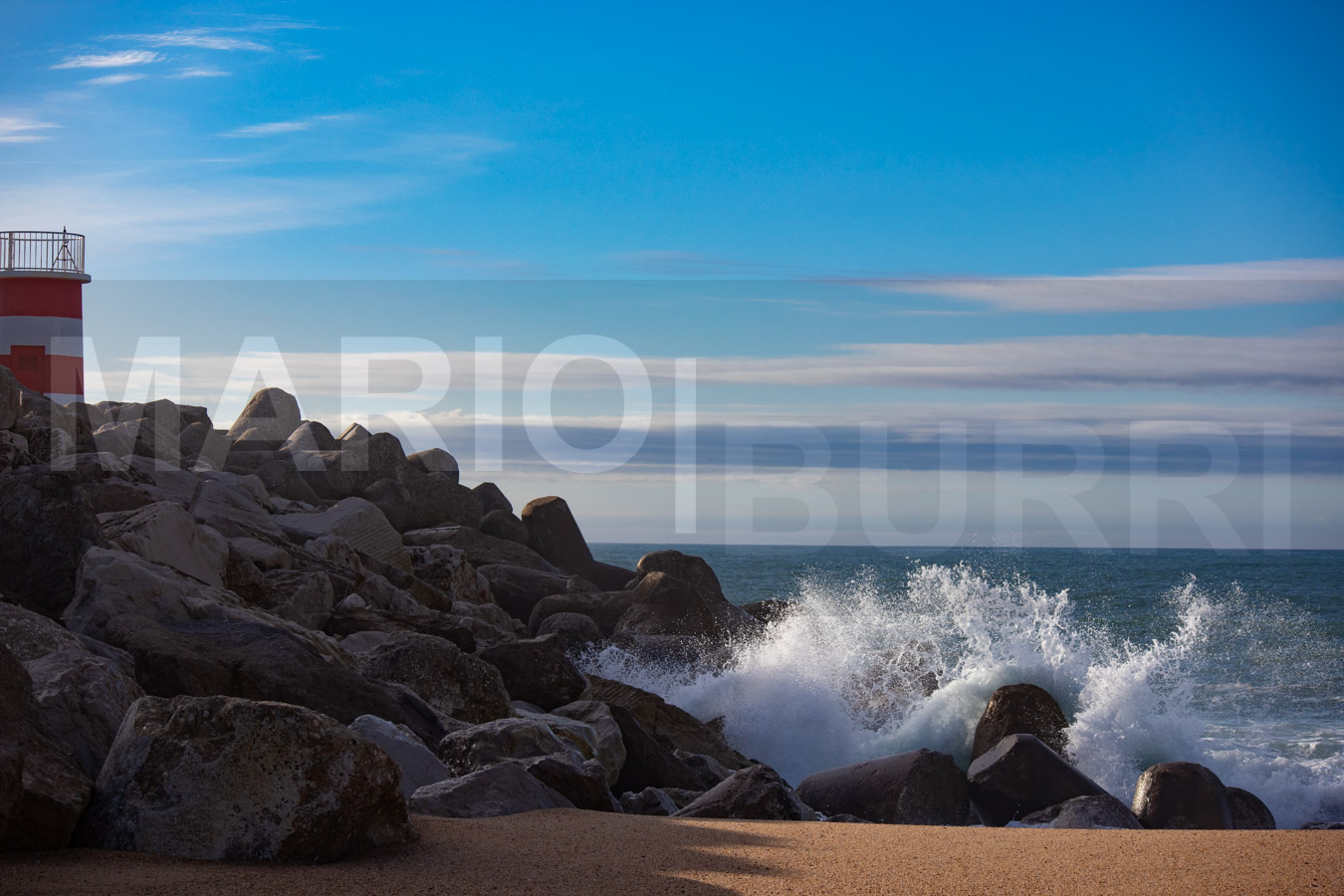 Ein sandiger Strandabschnitt mit grossen Felsen, an denen sich Wellen brechen, und einem rot-weiss gestreiften Leuchtturm auf der linken Seite unter einem klaren blauen Himmel. Foto von MARIO|BURRI.