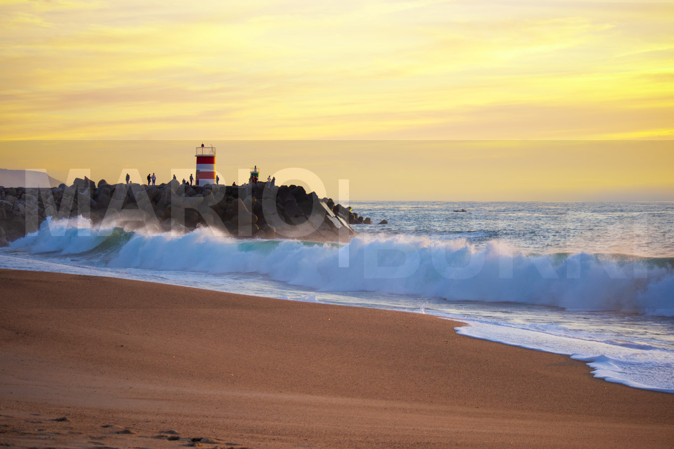Eine weite Ansicht des goldenen Sandstrandes von Nazaré, der sanft ins klare blaue Meer abfällt, mit dem charakteristischen Leuchtturm auf einer Klippe am Horizont unter einem klaren Himmel. Foto von MARIO|BURRI.