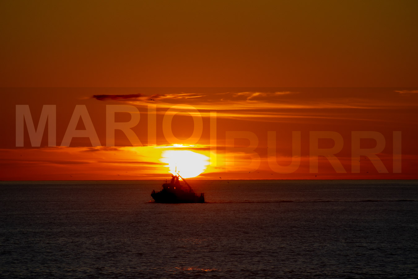 Ein Fischerboot fährt als dunkle Silhouette auf ruhigem Meer vor einem dramatischen Sonnenuntergangshimmel in warmen Orange- und Gelbtönen. Foto von MARIO|BURRI.